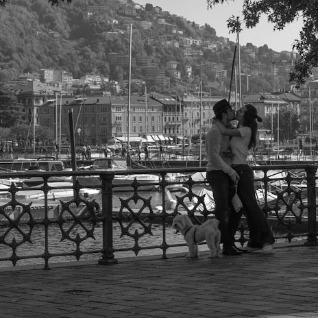 A couple embraces romantically by a waterfront promenade with a railing in the foreground. A small dog stands beside them. In the background, a marina with several boats is visible, along with hillside residential buildings. The scene has a tranquil and intimate atmosphere, enhanced by the grayscale color scheme.