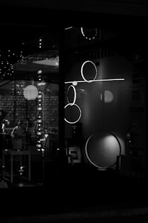 A black and white image of a storefront display featuring modern lighting fixtures, including circular and linear designs. The reflections and shadows add depth, creating a moody and sophisticated atmosphere.