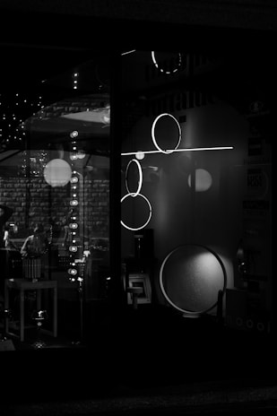 A black and white image of a storefront display featuring modern lighting fixtures, including circular and linear designs. The reflections and shadows add depth, creating a moody and sophisticated atmosphere.