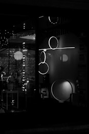 A black and white image of a storefront display featuring modern lighting fixtures, including circular and linear designs. The reflections and shadows add depth, creating a moody and sophisticated atmosphere.