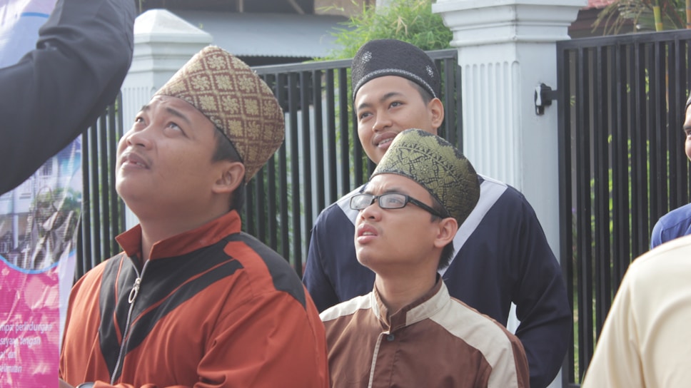 A group of men, possibly of Southeast Asian descent, are standing outdoors, dressed in traditional clothing with woven or patterned caps. They appear to be looking up at something above them with varying expressions of curiosity and interest. In the background, there is a black metal fence, some greenery, and parts of buildings.