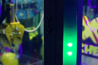 A close-up of a colorful amusement arcade claw machine, showcasing its mechanical gripping claw and vibrant lighting. The background includes blurred signage with neon colors, adding to the playful ambiance.