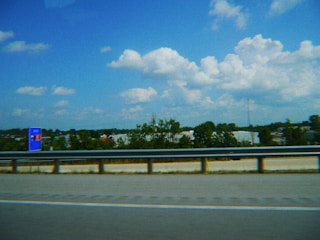 A highway scene featuring a metal guardrail in the foreground, with a clear blue sky and fluffy clouds in the background. Sparse greenery lines the roadside, and a blue and red sign stands on the left side near the road.
