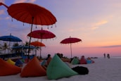 A serene beach scene with colorful umbrellas and bean bag chairs scattered on the sand. Several people are relaxing under the umbrellas, enjoying the vibrant sunset that paints the sky in shades of pink, purple, and orange. Palm trees are visible in the background near a building, adding to the tropical atmosphere.