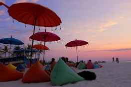 A serene beach scene with colorful umbrellas and bean bag chairs scattered on the sand. Several people are relaxing under the umbrellas, enjoying the vibrant sunset that paints the sky in shades of pink, purple, and orange. Palm trees are visible in the background near a building, adding to the tropical atmosphere.
