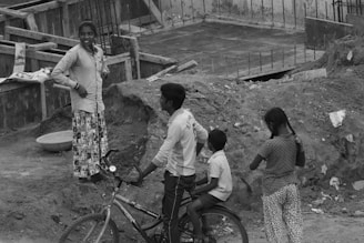 A group of children are gathered in a construction area. One child is sitting on a bicycle while another stands nearby, appearing to converse with a third child who is walking away. The area is surrounded by dirt, debris, and partially built structures.