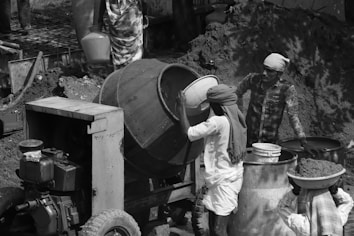 A construction site scene where three workers are engaged in mixing and preparing construction materials. One worker is operating a cement mixer while another holds a bucket, ready to pour its contents. The third person is carrying a large bowl on their head, filled with mixed cement or soil. They are surrounded by tools and machinery, and dressed in traditional work attire with head coverings.