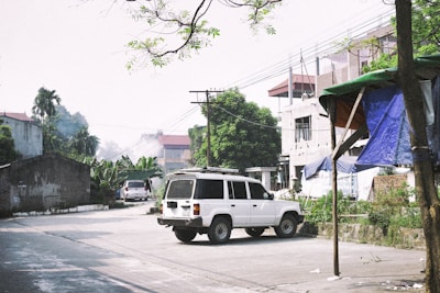 A white SUV is parked on a narrow street lined with various types of buildings, including an incomplete structure and a small brick building. Overhead wires and utility poles are visible, while trees and leafy plants, such as banana trees, add greenery to the surroundings. A canopy made of blue and green tarps is set up near the sidewalk.