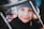 Young child wearing a grey beanie, smiling warmly at the camera with a hint of playfulness. The background is slightly blurred, with another child partially visible, creating a candid and casual atmosphere. The focus is on the child's expressive eyes and charming dimples.