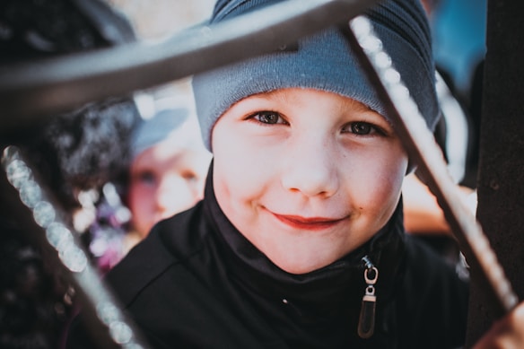 Young child wearing a grey beanie, smiling warmly at the camera with a hint of playfulness. The background is slightly blurred, with another child partially visible, creating a candid and casual atmosphere. The focus is on the child's expressive eyes and charming dimples.