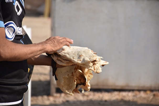 A person is holding several pieces of flatbread, possibly freshly made. The background is out of focus, with a neutral tone. The person's hand and part of the arm are visible, wearing a dark shirt with some logos or designs.