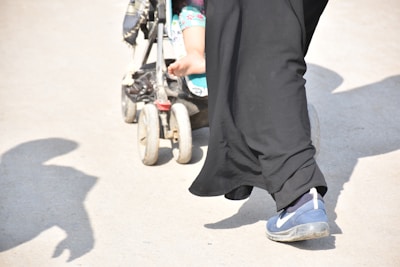 A person wearing black clothing and blue sneakers is pushing a stroller on a concrete surface. Part of a child's leg with floral patterns on their trousers is visible in the stroller. Shadows are cast on the ground, suggesting bright sunlight.