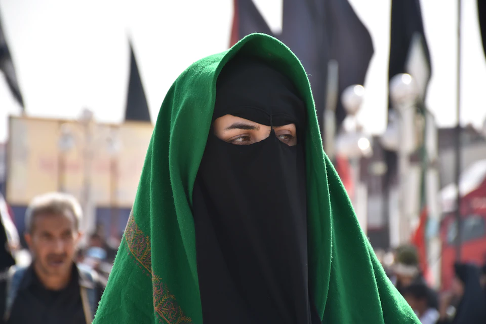 The image shows a person wearing a green headscarf and black burka, also known as a hijab or niqab, with their eyes closed. The individual is standing against a backdrop of several flags, suggesting they may be in an outdoor event or gathering. In the background, there are other people present, although they are blurred out and not the main focus of the image.
The person's attire and the presence of multiple flags create a sense of depth and interest within the scene.