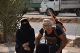 Two people are sitting outdoors near a road, one wearing a black niqab and the other in casual clothing with a traditional keffiyeh. A large backpack with colorful ribbons is visible, and a truck is parked in the background near a small palm tree.