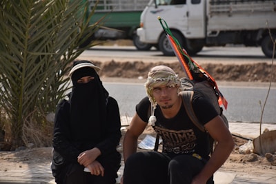 Two people are sitting outdoors near a road, one wearing a black niqab and the other in casual clothing with a traditional keffiyeh. A large backpack with colorful ribbons is visible, and a truck is parked in the background near a small palm tree.