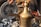 A brass coffee pot with an ornate lid is positioned among charcoal pieces. A hand holds a soot-covered metal plate in the background, adding to the rustic, traditional setting. Various jars and containers are partially visible, suggesting a market or outdoor setting.