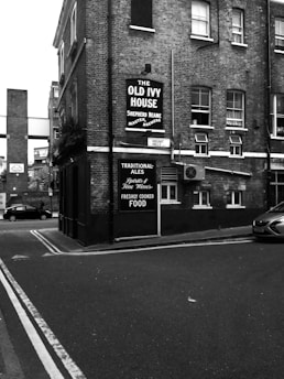 A street scene featuring a corner building with signage for 'The Old Ivy House' pub, advertising traditional ales and freshly cooked food. The architecture is characterized by brick walls and multiple windows. A car is parked along the side of the street, with another vehicle visible in the background.