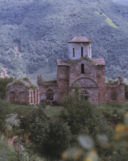 An ancient, stone-built church stands amidst dense greenery and mountainous terrain. The structure shows signs of age and partial ruin, with its dome intact but walls overgrown with vegetation. The background is a lush, forested mountainside, creating a serene and remote setting.