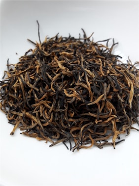 A pile of dried tea leaves featuring long, curly strands is placed on a white surface, exhibiting a mix of dark and golden hues.
