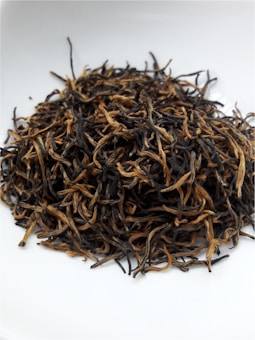 A pile of dried tea leaves featuring long, curly strands is placed on a white surface, exhibiting a mix of dark and golden hues.