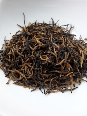 A pile of dried tea leaves featuring long, curly strands is placed on a white surface, exhibiting a mix of dark and golden hues.