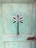 A weathered wooden door painted in a faded turquoise color. The door features a decorative cut-out pattern resembling a flower or star shape. There is visible wear and tear, with some cracks and chipped paint. The door has a rusty, metal latch.