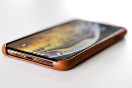 A smartphone lies flat on a surface, partially encased in a brown protective cover. The device's screen is on, displaying a swirling pattern of warm colors like yellow and orange. The phone is positioned at an angle showing the speaker grille and lightning port clearly.