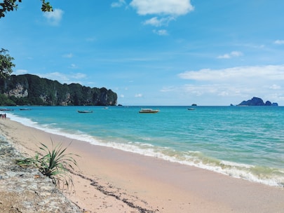 A serene beach scene with turquoise waters gently lapping against a sandy shore. Longtail boats float peacefully on the water. The horizon is dotted with distant islands under a clear sky. Lush green cliffs rise on one side, and there are a few people walking along the shoreline.
