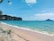 A serene beach scene with turquoise waters gently lapping against a sandy shore. Longtail boats float peacefully on the water. The horizon is dotted with distant islands under a clear sky. Lush green cliffs rise on one side, and there are a few people walking along the shoreline.