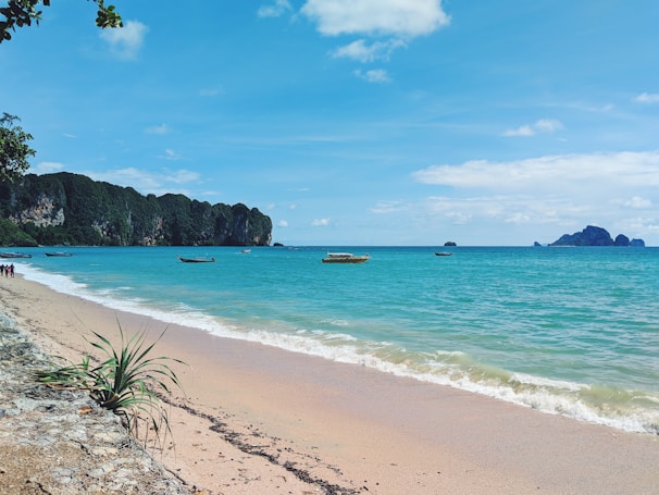 A serene beach scene with turquoise waters gently lapping against a sandy shore. Longtail boats float peacefully on the water. The horizon is dotted with distant islands under a clear sky. Lush green cliffs rise on one side, and there are a few people walking along the shoreline.