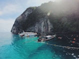 A picturesque coastal scene with clear turquoise water, a towering cliff with dense greenery on top, and several boats floating on the water. People are swimming and snorkeling around the boats, suggesting a recreational activity in a serene natural setting under the sunlight.