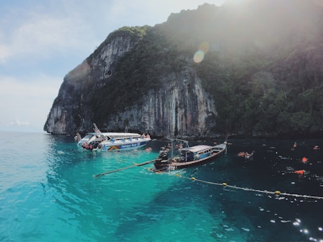 A picturesque coastal scene with clear turquoise water, a towering cliff with dense greenery on top, and several boats floating on the water. People are swimming and snorkeling around the boats, suggesting a recreational activity in a serene natural setting under the sunlight.