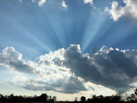 Sunshine breaks through fluffy clouds creating striking rays of sunlight stretching across a blue sky. The scene is set above a landscape with silhouettes of trees along the horizon.