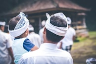 A man carrying a child on his shoulder, both wearing traditional white headpieces with intricate patterns. They appear to be part of a larger group, possibly at a cultural or ceremonial gathering. The environment around them is out of focus, suggesting a lush or temple-like setting.
