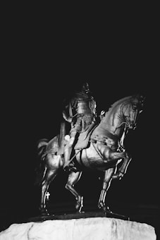 A black and white statue of a warrior in armor riding a horse, poised on a pedestal against a dark background.