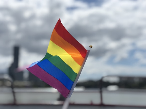 A rainbow-colored flag is prominently displayed against a cloudy sky, with a slight blur of urban structures in the background. The flag is rippling in the gentle breeze, emphasizing the vibrant colors.
