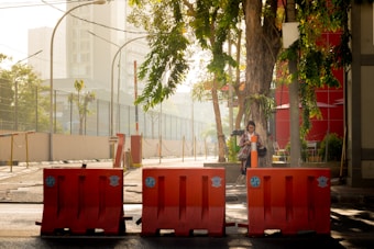 A quiet urban street scene with bright orange barricades in the foreground and a large tree providing shade. A person stands near the tree holding a digital device. The sun casts a warm glow over the area, with buildings and greenery visible in the background.