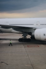 A large aircraft is parked on an airport tarmac with its landing gear visible. A person wearing a reflective safety vest is walking near the airplane. The sky appears overcast with clouds.