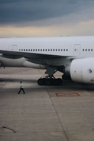 A large aircraft is parked on an airport tarmac with its landing gear visible. A person wearing a reflective safety vest is walking near the airplane. The sky appears overcast with clouds.