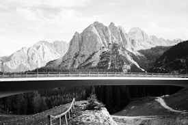 A breathtaking mountain range stretches across the background with sharp, jagged peaks. Below, a modern bridge spans the width of the image, elevated and supported by pillars. The foreground features a dirt path with a railing and sparse vegetation. The entire scene is captured in black and white, emphasizing the contrast between the rugged natural landscape and the sleek architectural structure.