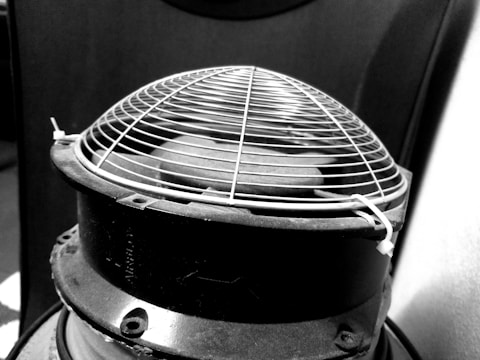 A close-up of a mechanical object with a protective wire mesh cover, likely part of an industrial or electronic device. The structure is cylindrical with visible screws and a tie securing the mesh.