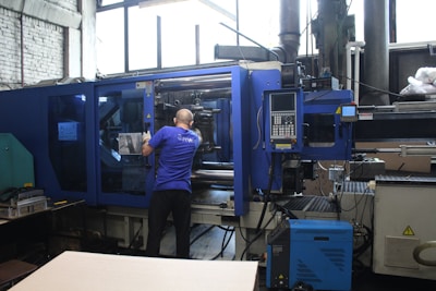 A person is operating a large industrial machine in a factory setting. The machine has a blue exterior with a control panel featuring numerous buttons and a screen. The factory environment includes visible pipes, control boxes, and other equipment.