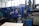 A person is operating a large industrial machine in a factory setting. The machine has a blue exterior with a control panel featuring numerous buttons and a screen. The factory environment includes visible pipes, control boxes, and other equipment.