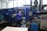 A person is operating a large industrial machine in a factory setting. The machine has a blue exterior with a control panel featuring numerous buttons and a screen. The factory environment includes visible pipes, control boxes, and other equipment.