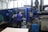 A person is operating a large industrial machine in a factory setting. The machine has a blue exterior with a control panel featuring numerous buttons and a screen. The factory environment includes visible pipes, control boxes, and other equipment.