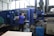 A person is operating a large industrial machine in a factory setting. The machine has a blue exterior with a control panel featuring numerous buttons and a screen. The factory environment includes visible pipes, control boxes, and other equipment.