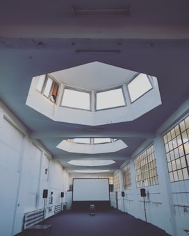 An industrial-style room with high ceilings and large octagonal skylights, allowing natural light to filter in. White walls and square windows create a minimalistic atmosphere. A large screen is set up at one end, surrounded by speakers and other equipment, suggesting a space for presentations or viewings.
