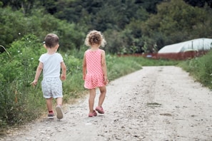Two young children are walking down a dirt path surrounded by lush green vegetation. The girl, wearing a pink polka-dot dress, and the boy, in a white shirt and patterned shorts, are heading towards a garden area. In the background, there's a greenhouse, and the area is enclosed by a wooden fence.