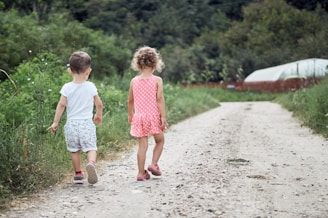 Two young children are walking down a dirt path surrounded by lush green vegetation. The girl, wearing a pink polka-dot dress, and the boy, in a white shirt and patterned shorts, are heading towards a garden area. In the background, there's a greenhouse, and the area is enclosed by a wooden fence.