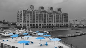 A grayscale cityscape features a large, historic brick building with intricate architectural details facing a waterfront. In the foreground, there is a sandy beach area with bright blue umbrellas and chairs that stand out against the monochrome background. Several people are relaxing on the beach. To the left, a marina is visible with numerous boats docked along the water.